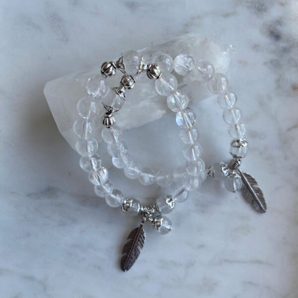 clear quartz beaded bracelet with silver feather charm on a marble surface